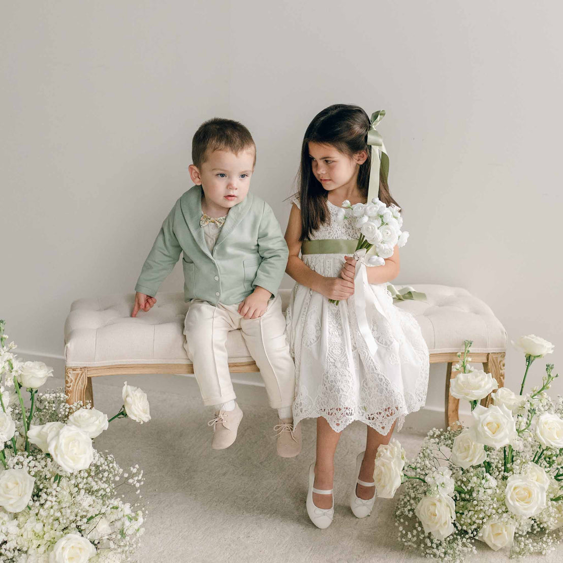 A toddler wearing the Ezra Sage 4-Piece Suit sits on a bench next to a girl holding a white floral bouquet.