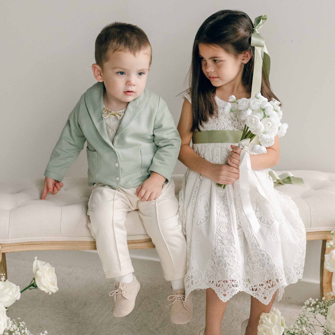 A toddler wearing the Ezra Sage 4-Piece Suit sits on a bench next to a girl holding a white floral bouquet.
