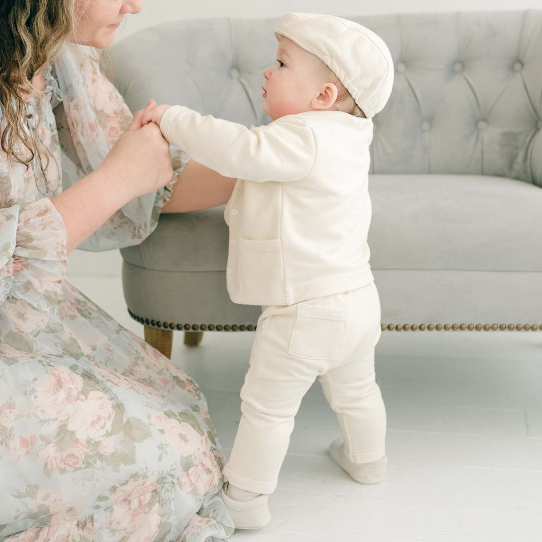 A baby in the Ezra Tan 4-Piece Suit and cap, holding onto a woman's hands for support.
