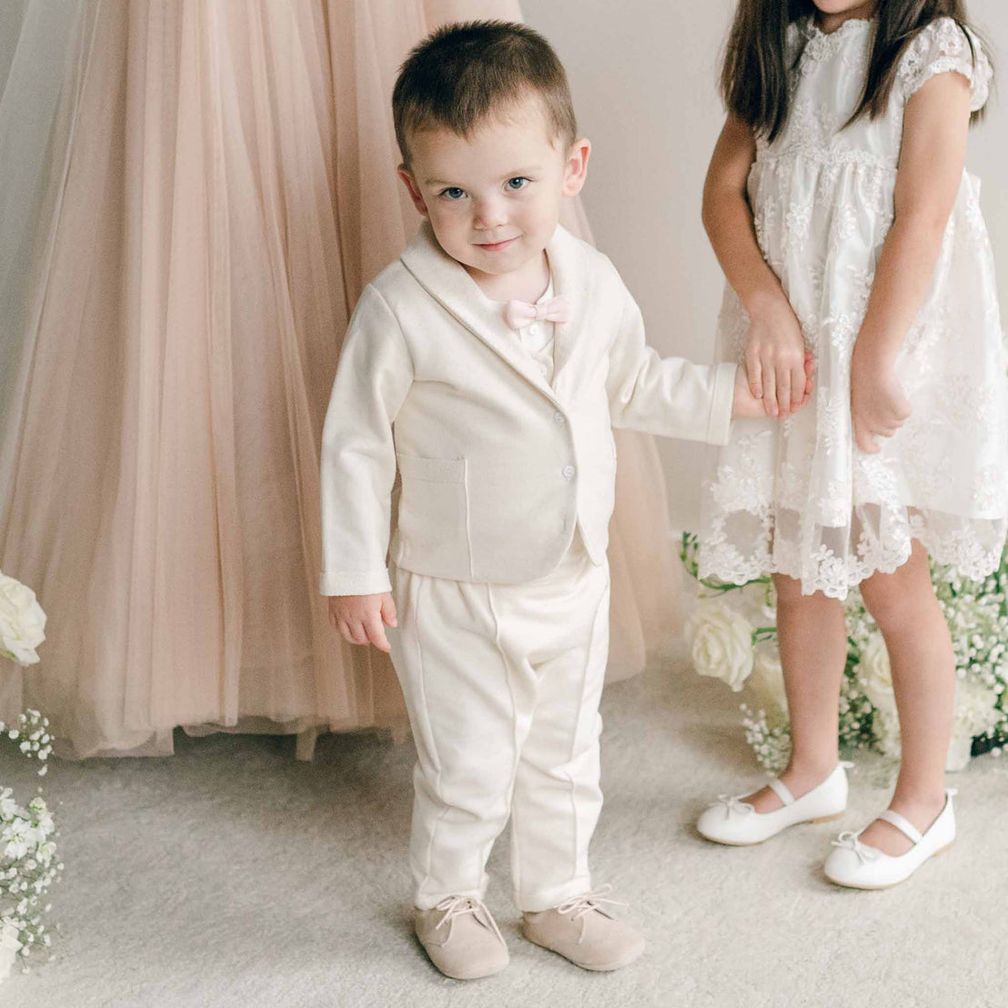 A toddler wearing the Ezra Tan 4-Piece Suit, standing and holding hands with a girl in a lace dress.