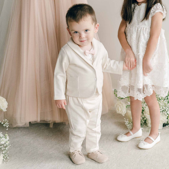 A toddler wearing the Ezra Tan 4-Piece Suit, standing and holding hands with a girl in a lace dress.
