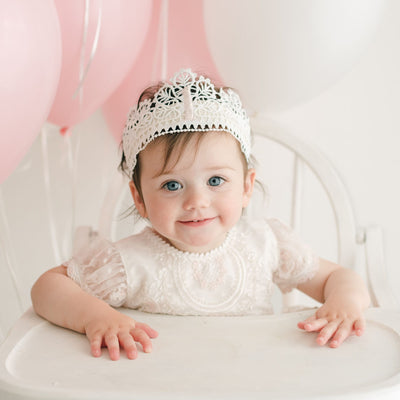 A toddler wearing a lace dress and the First Birthday Crown sits in a white high chair. Pink and white balloons are tied to the chair. She looks at the camera with a smile, her hand resting on the high chair tray in front of her.