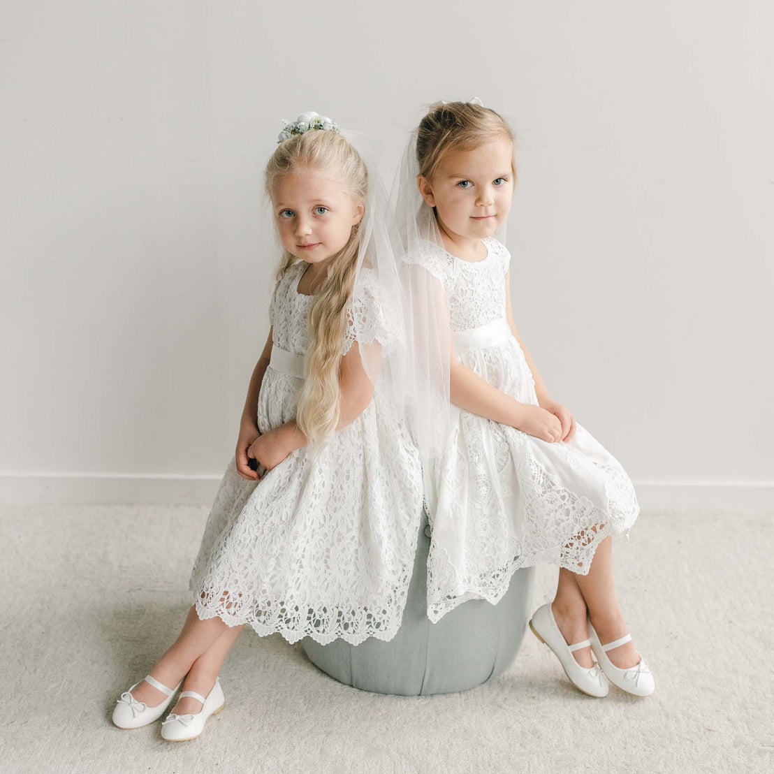Two girls in matching lace dresses and satin sashes sit together, one wearing a white floral veil and the other a bow with a flowing veil.