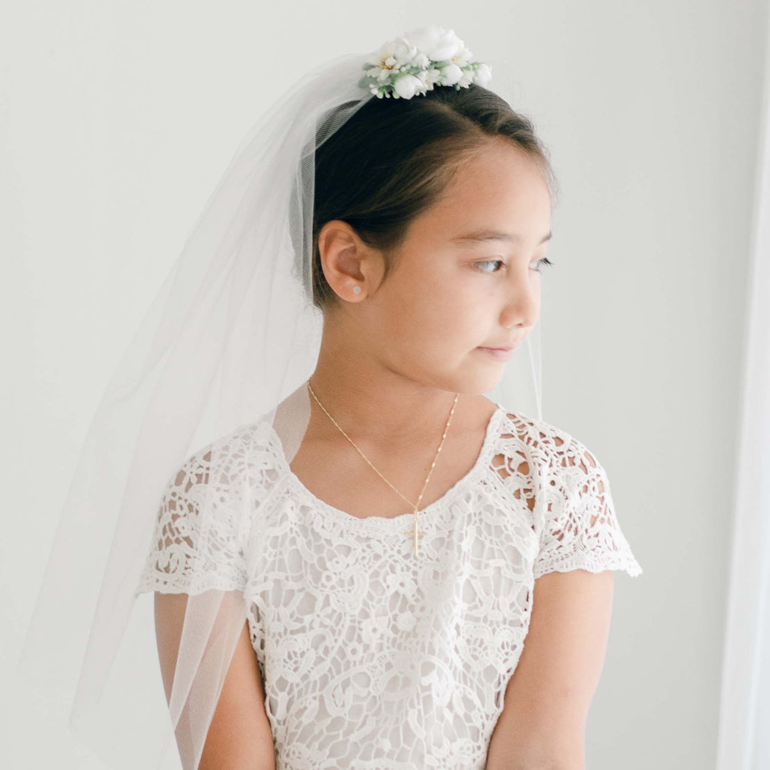 A girl in a lace dress gazes to the side, wearing a white floral hairpiece and tulle veil secured at the crown of her head.