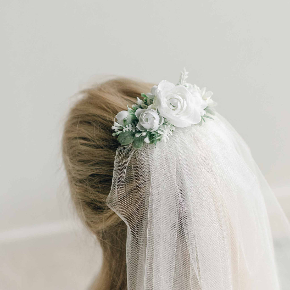 Close-up of a white floral hairpiece with soft green accents, attached to a tulle veil placed on top of a girl's hairstyle.