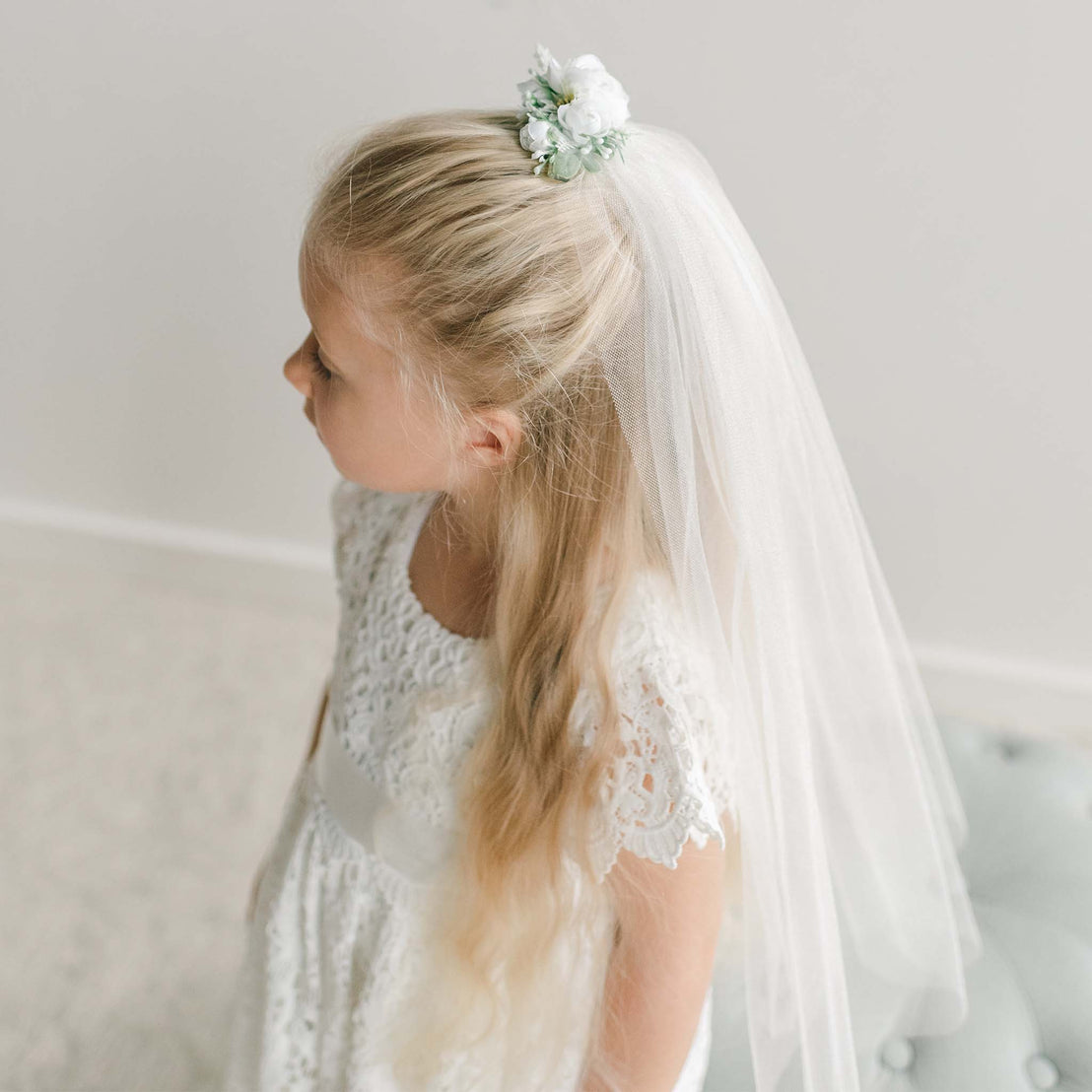 A young girl wearing a lace dress with a satin sash, styled with a white floral hairpiece and delicate tulle veil cascading down her back.