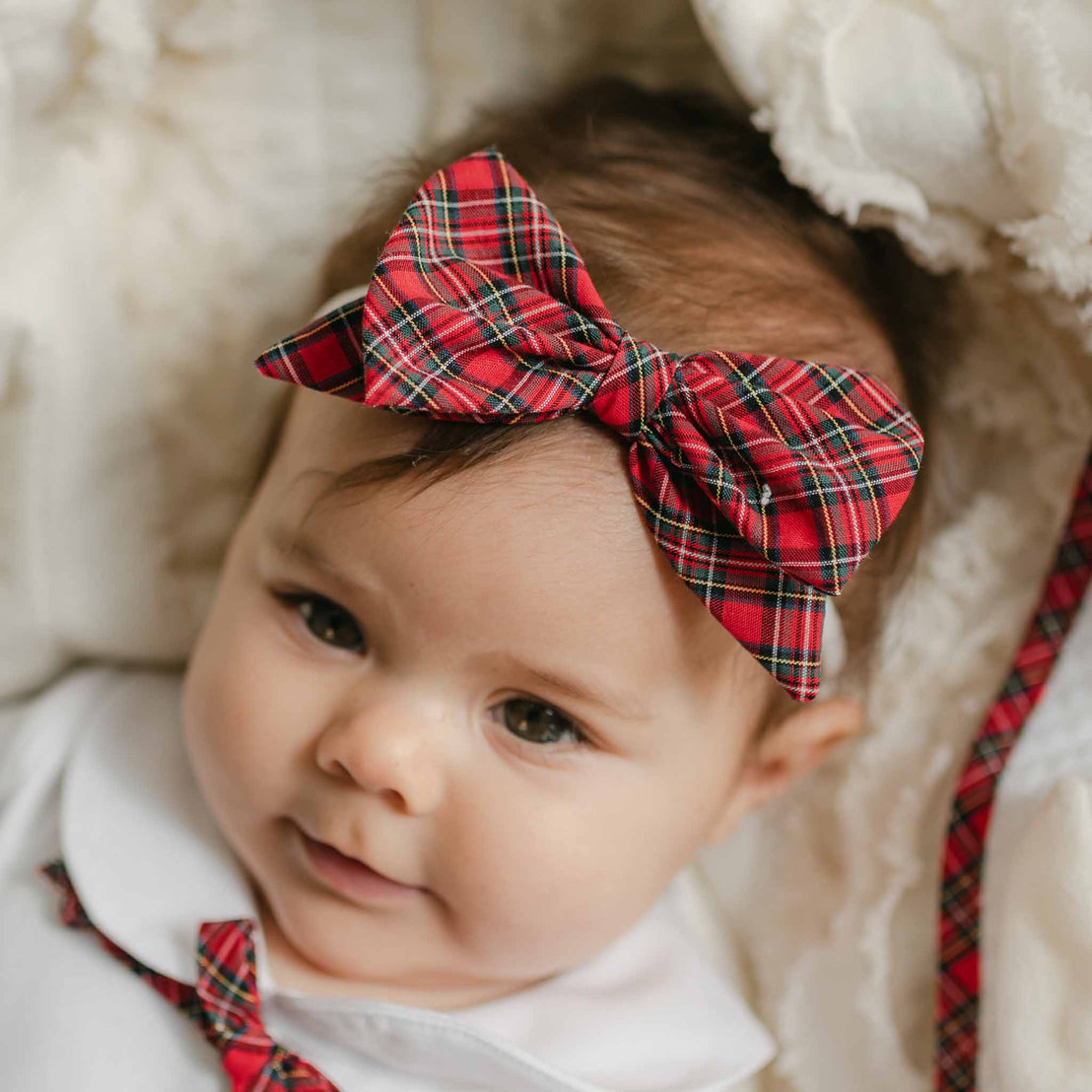 A baby with dark hair is wearing a handcrafted Gabriella Bow Headband in red plaid, along with a white outfit featuring a matching plaid tie. The baby lies on a soft, light-colored blanket, looking content and cozy.