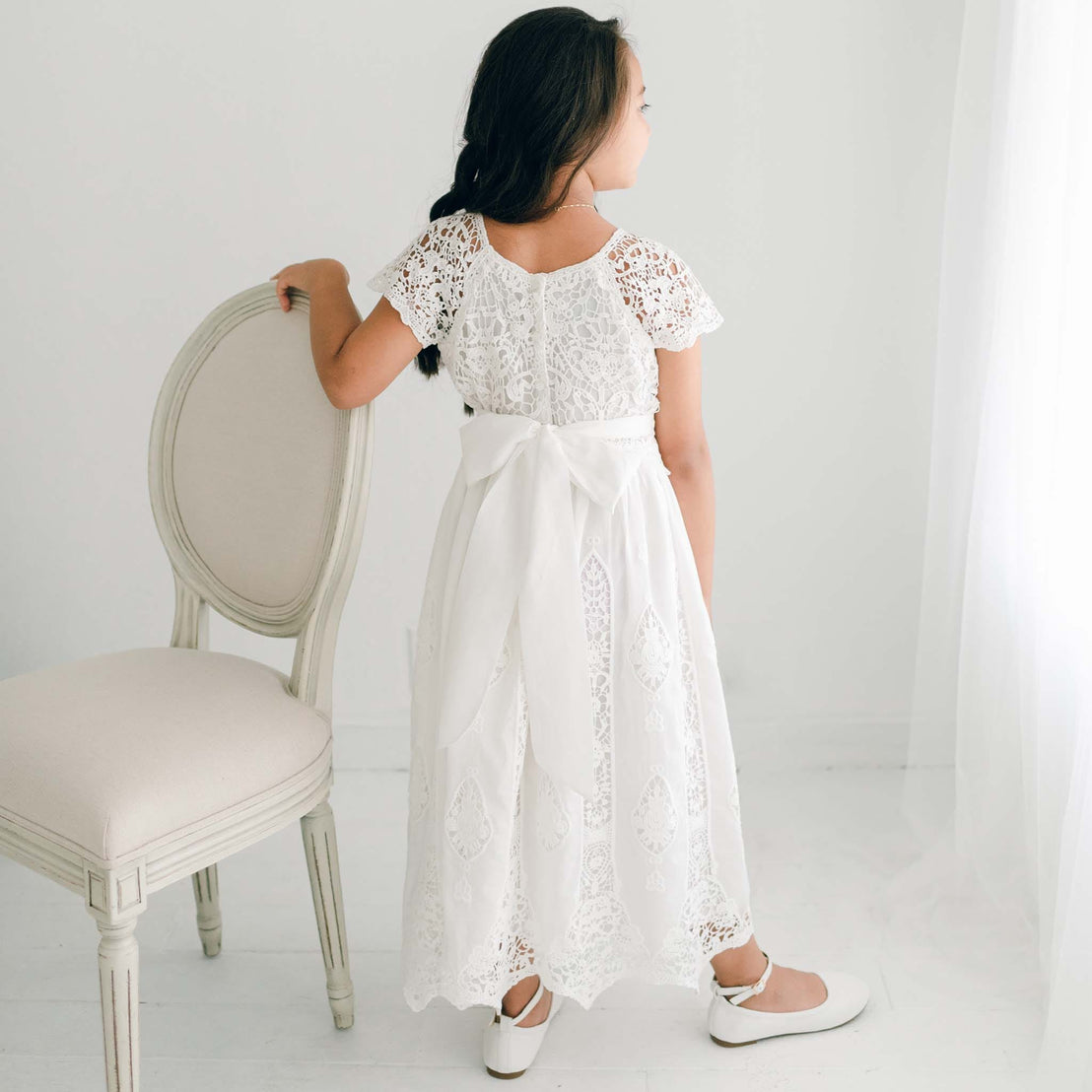 Back view of a girl wearing the Grace Girls Lace Dress with an all-white sash tied in a large bow.