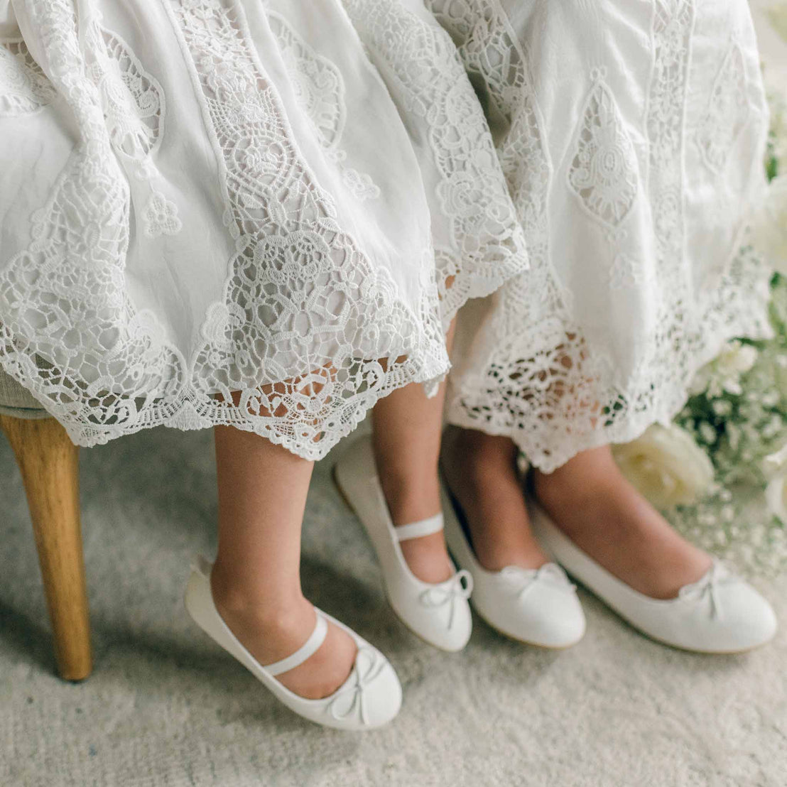 Close-up of two girls' feet wearing white shoes with bows, showing the scalloped lace hem of the Grace Girls Lace Dress.