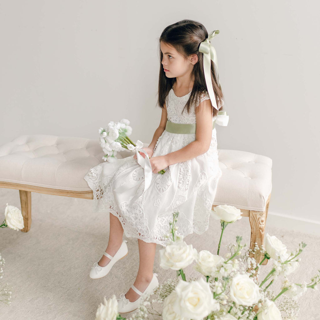 A girl sitting on a bench wearing the Grace Girls Lace Dress with a light green sash and matching hair bow, holding a white bouquet.