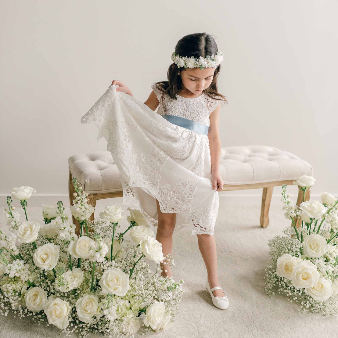 A girl wearing the Grace Girls Lace Dress with a light blue sash, lifting the side of her skirt while standing among white roses.