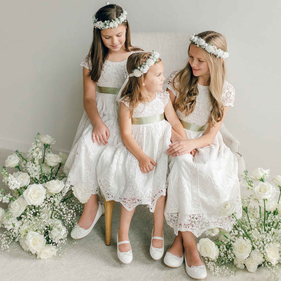 Three girls wearing the Grace Girls Lace Dress styled with light green sashes and white flower wreaths, seated among white roses.