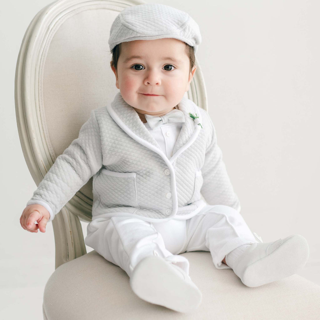 Baby wearing the Grayson 3-Piece Suit with quilted jacket, white pants, matching cap, and bow tie seated on a light chair.
