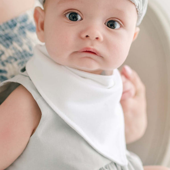 A baby dressed in a light grey French Terry cotton outfit with a white Grayson Bandana Bib, looking directly at the camera. The baby is seated on an adult's lap whose face is not visible. The background is softly blurred, focusing attention on the baby.