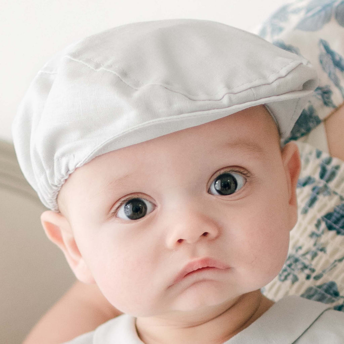A baby wearing the Grayson Linen Newsboy Cap stares ahead with large, expressive eyes. Dressed in the matching Linen Romper. A partial view of another person is visible in the background. The baby’s expression appears curious and intrigued.