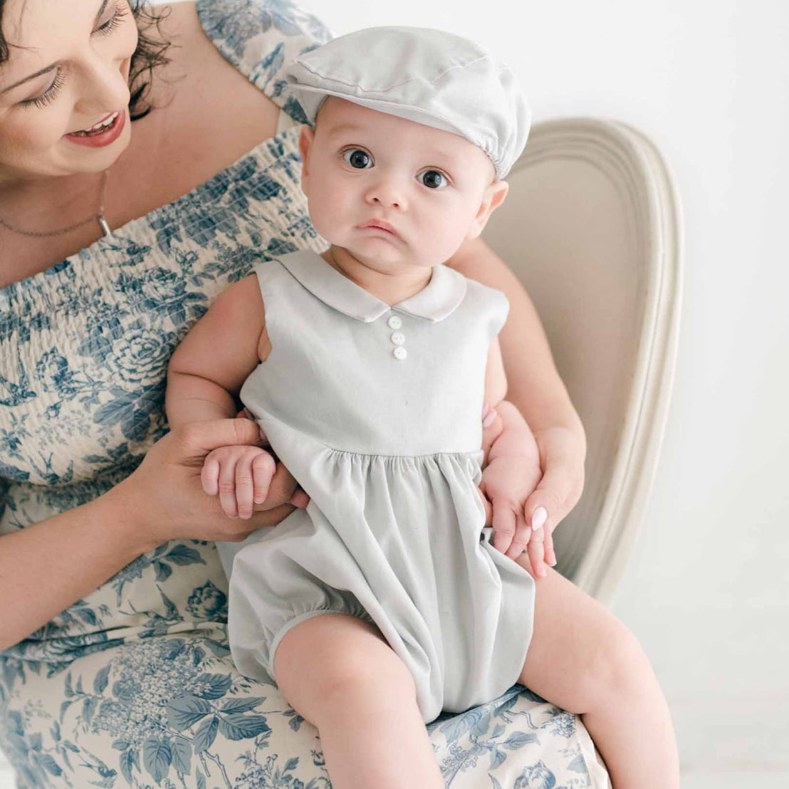 A woman in a blue floral dress sits on a chair, holding a baby dressed in the Grayson Linen Romper and matching Newsboy Cap. The baby, with a curious expression, is perched on the woman's lap, and the background is softly lit with a neutral-toned setting.