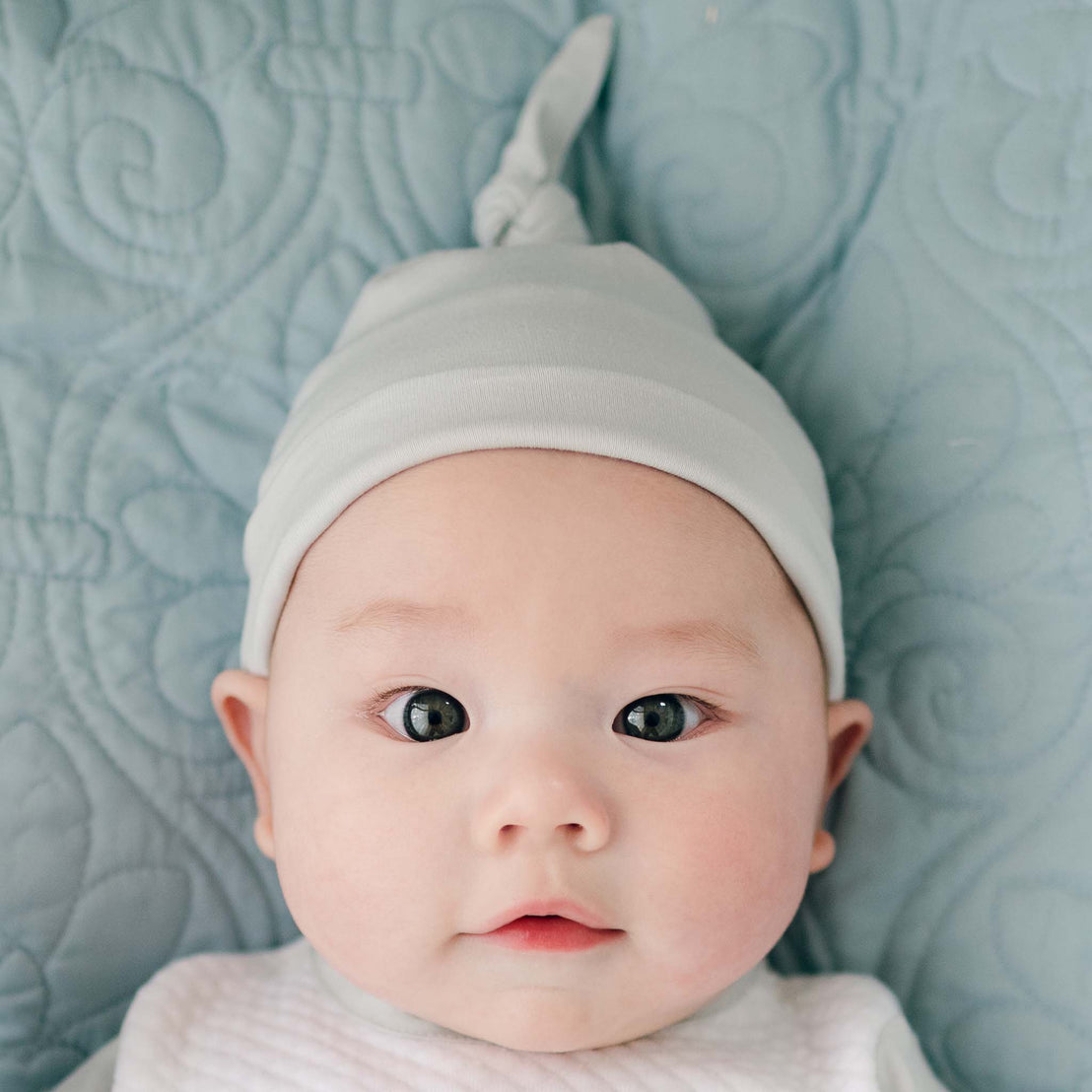 A baby in a Grayson Newborn Knot Cap, a light green hat with a stylish knot, lies on a quilted blue blanket. With wide eyes and slightly parted lips, the infant gazes directly at the camera, exuding charm and innocence.