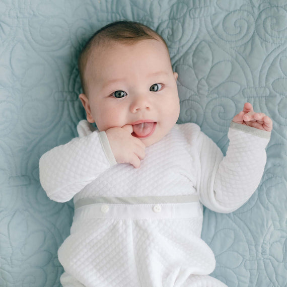 A baby wearing a designer take home outfit lies on a light blue quilted blanket. The newborn, dressed in a Grayson Quilted Romper, gazes at the camera with wide eyes, their tongue sticking out slightly as their right hand nears their mouth.