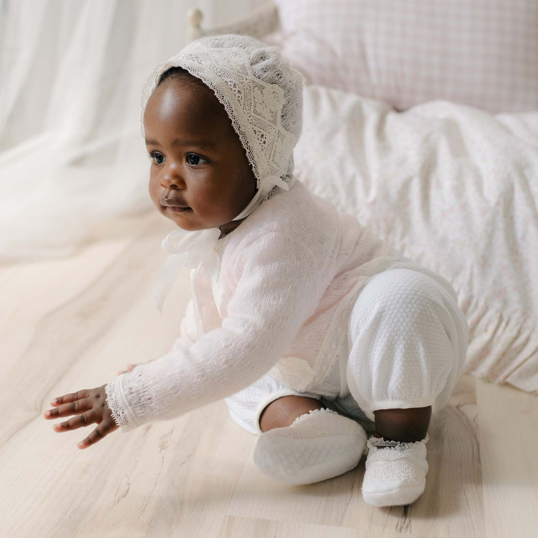 Baby in the Hailey Knit Bonnet and matching sweater romper sitting on a wood floor. Details show the intricate light ivory lace on the bonnet. A perfect baptism gift from godparents.
