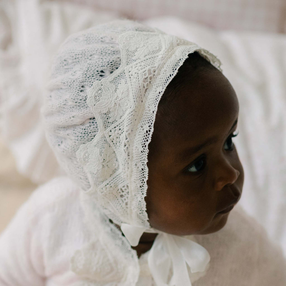 Close up of a baby wearing the Hailey Knit Bonnet against a soft, blurred background. Details show the knit and matching light ivory laces. 