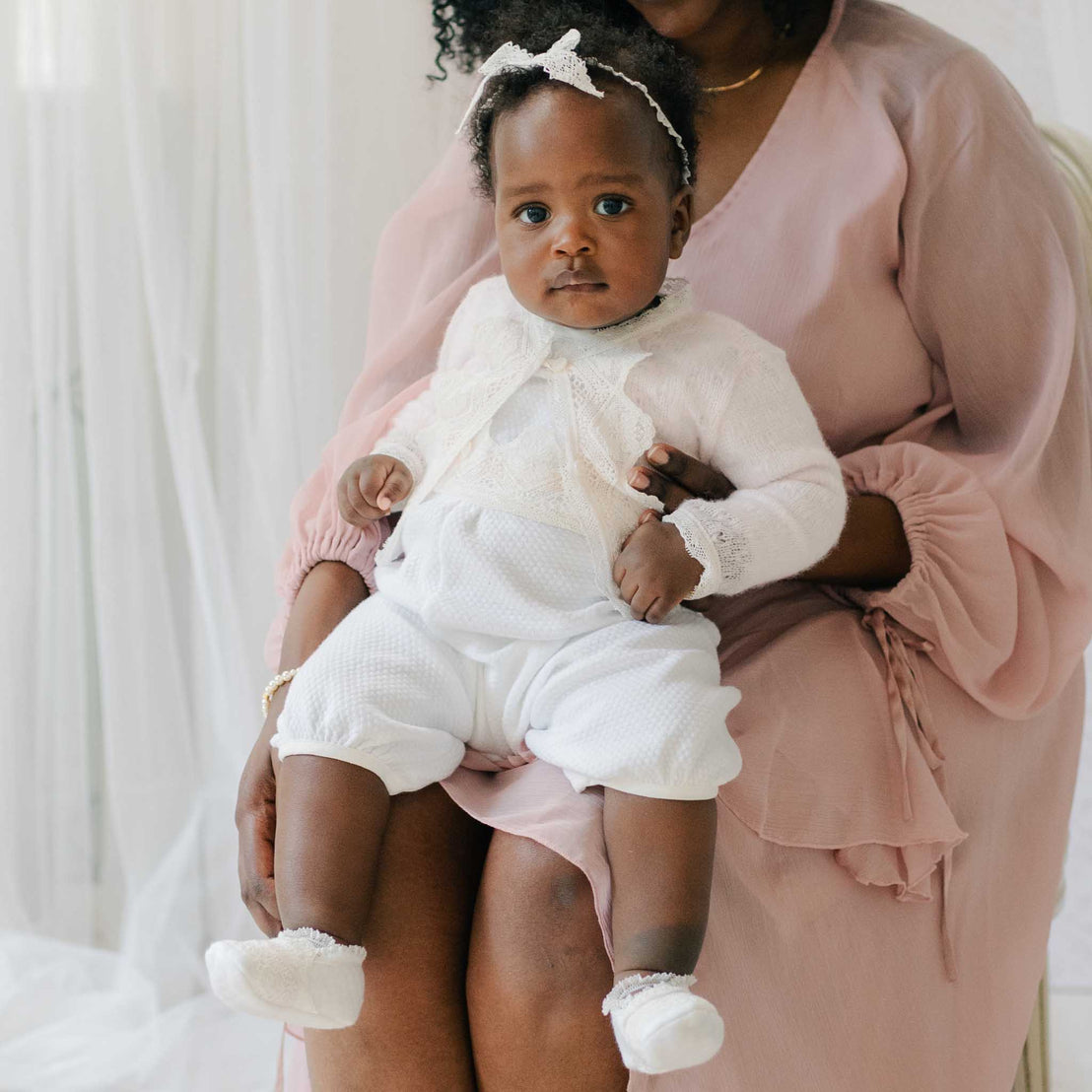 Baby wearing the Hailey Romper and matching Lace Headband. She is gently held by a woman in a pink dress against a white sheer background.