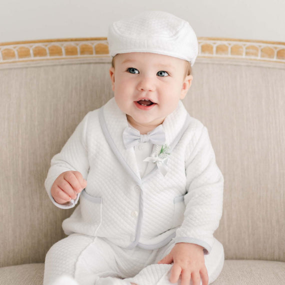 Baby sitting on a cushioned bench wearing the Harrison 3-Piece Pants Suit, featuring a quilted jacket with soft blue trim, bow tie, and matching textured cap.