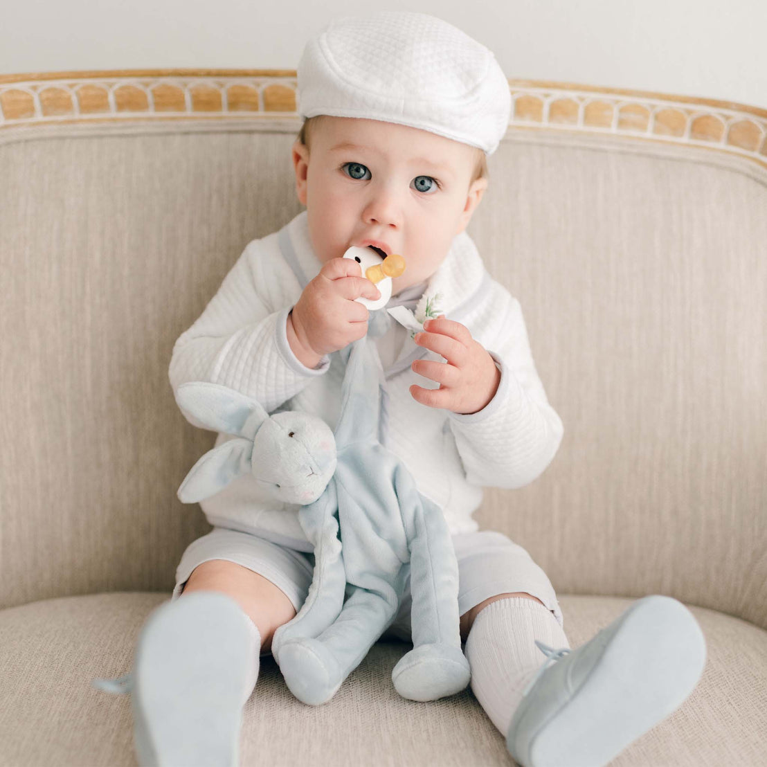 A baby in the Harrison 3-Piece Shorts Suit sits on a beige chair, holding a plush blue bunny and a pacifier while wearing the quilted white jacket, bow tie, and shorts.