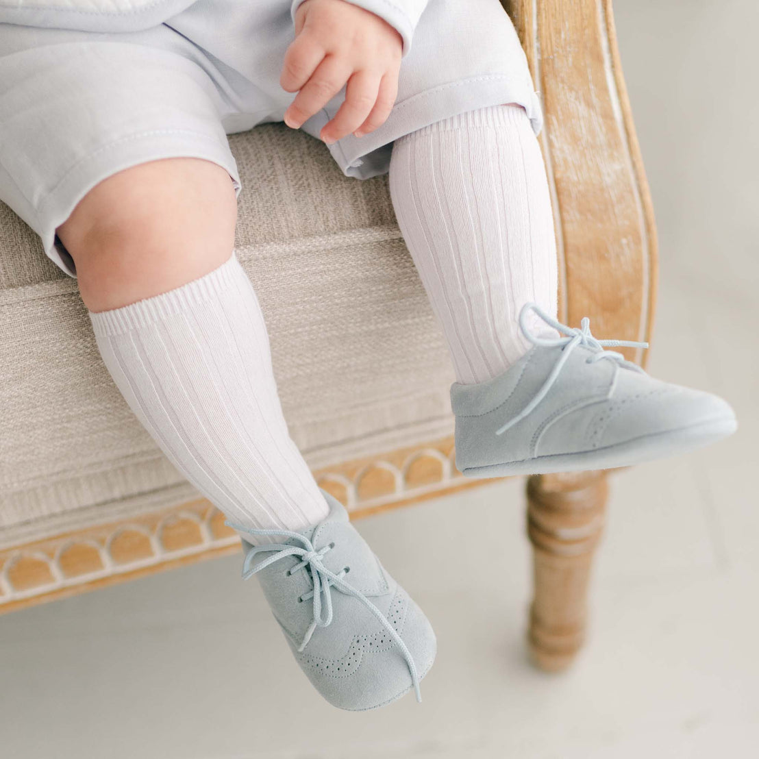  A close-up of a baby’s legs wearing the Harrison 3-Piece Shorts Suit, showcasing ribbed white knee-high socks and soft blue suede booties with lace-up detail.