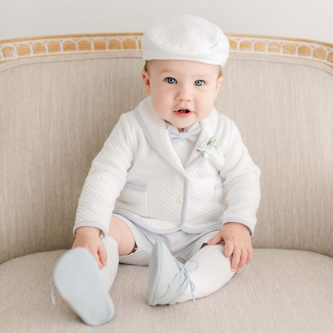 A baby sits on a beige upholstered chair wearing the Harrison 3-Piece Shorts Suit, featuring a quilted white jacket with light blue trim, matching shorts, and a coordinating newsboy cap.
