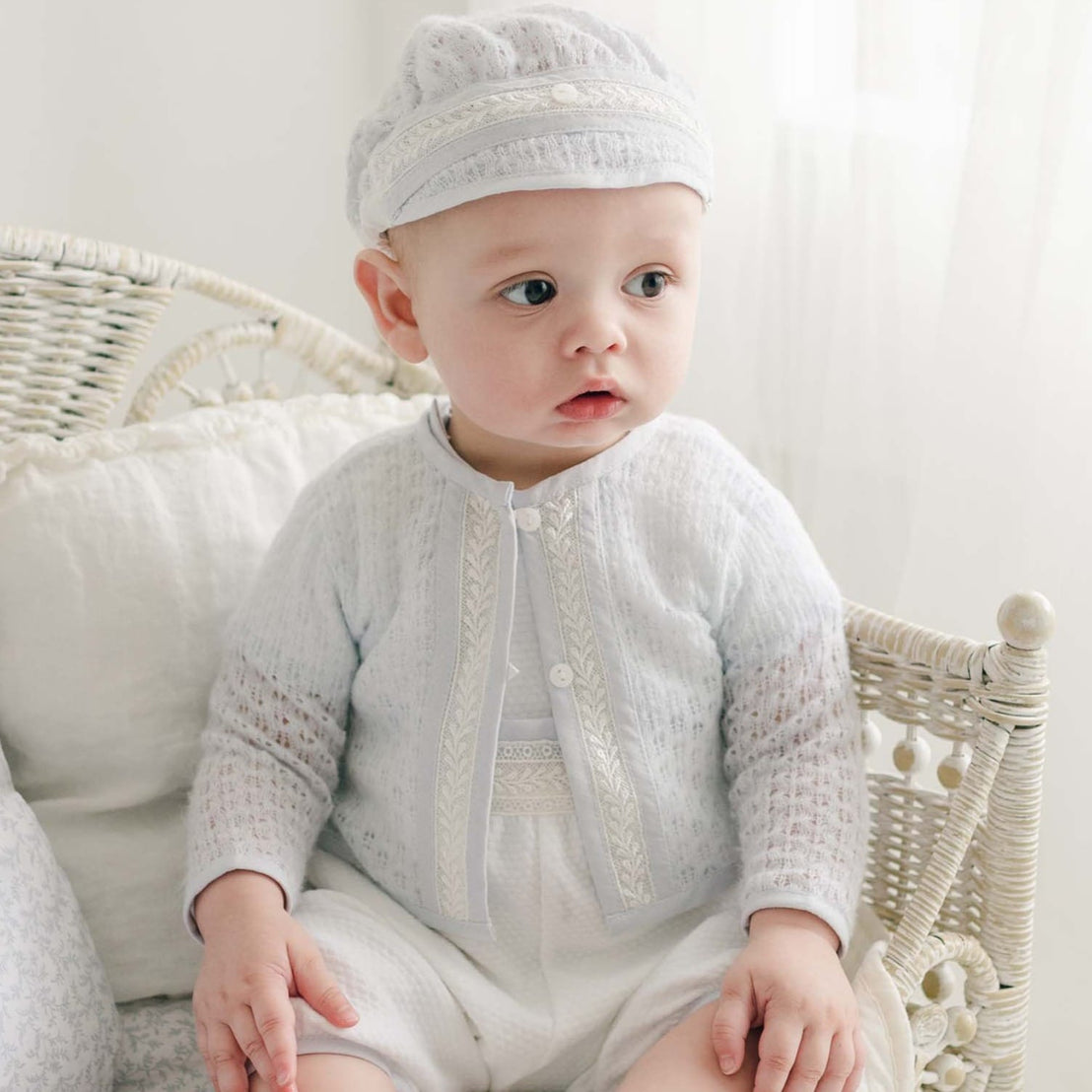 Baby boy seated on a wicker chair, dressed in a delicate blue knit sweater with ivory lace detailing and white quilted pants.