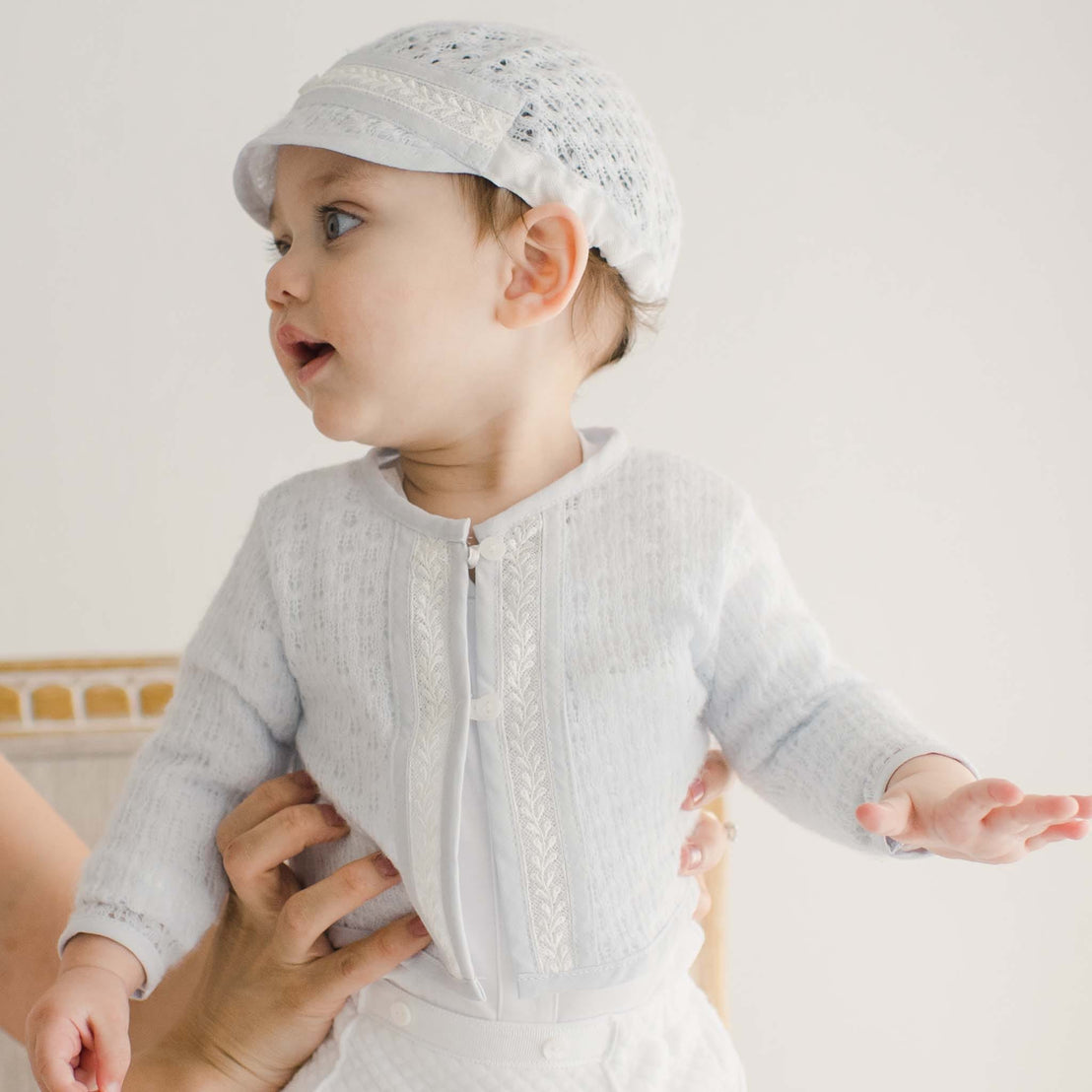 Baby boy held upright, wearing a soft blue knit sweater with lace accents, paired with white quilted pants.