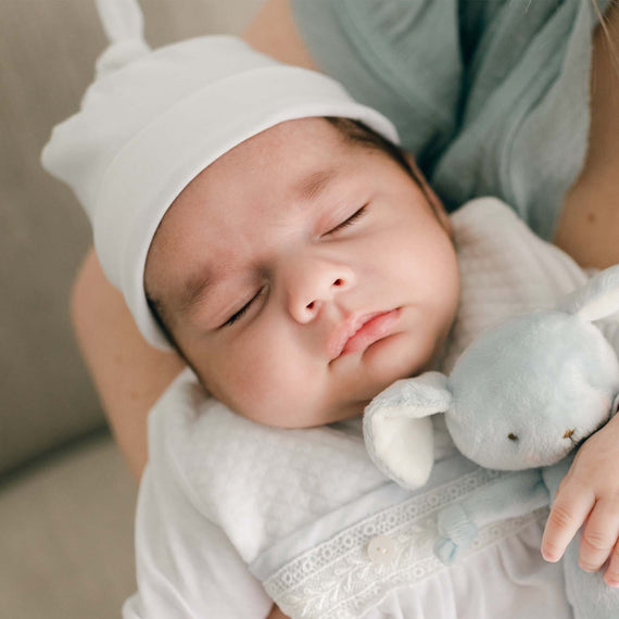 A sleeping baby wearing the Harrison Knot Cap, a soft white cotton hat with a knotted top and folded brim.