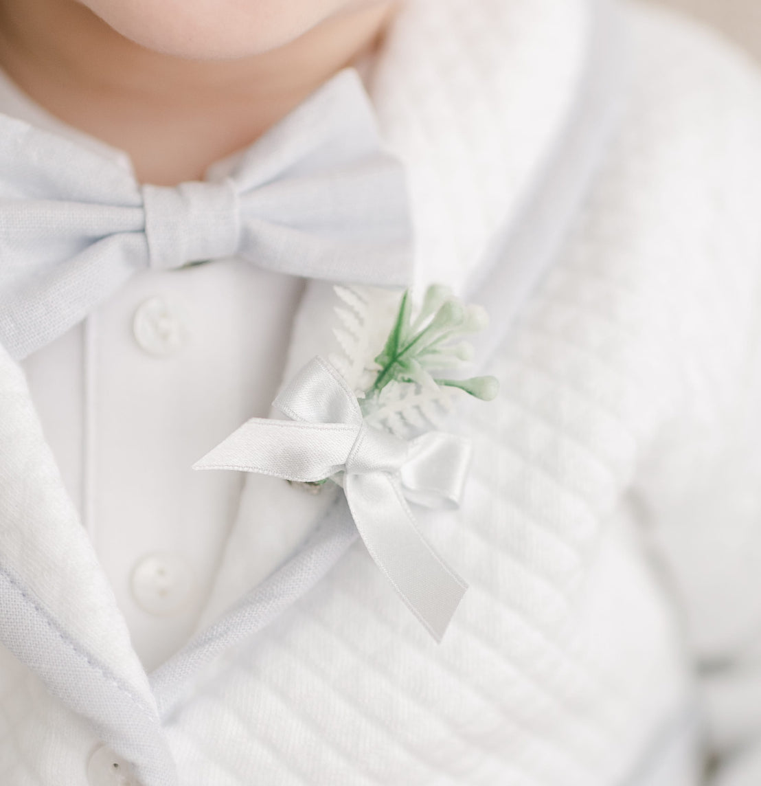 Close-up of the Harrison Linen Bow Tie & Boutonniere, highlighting the soft blue linen bow tie and elegant boutonniere with white ribbon and green accents, styled on a quilted white jacket.