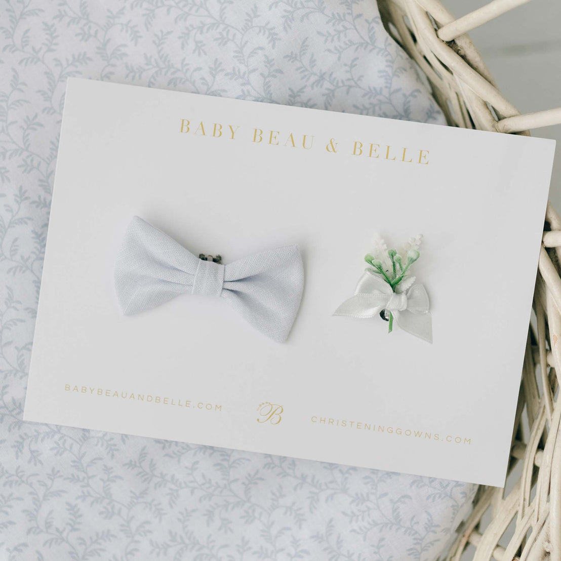 A flat lay display of a pale blue linen bow tie and a delicate boutonniere with white ribbon and green accents, showcased on a branded Baby Beau & Belle card.