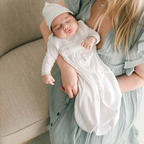 A newborn baby sleeping peacefully in a parent's arms, dressed in the Harrison Newborn Gown with textured white fabric and delicate lace details at the waist.