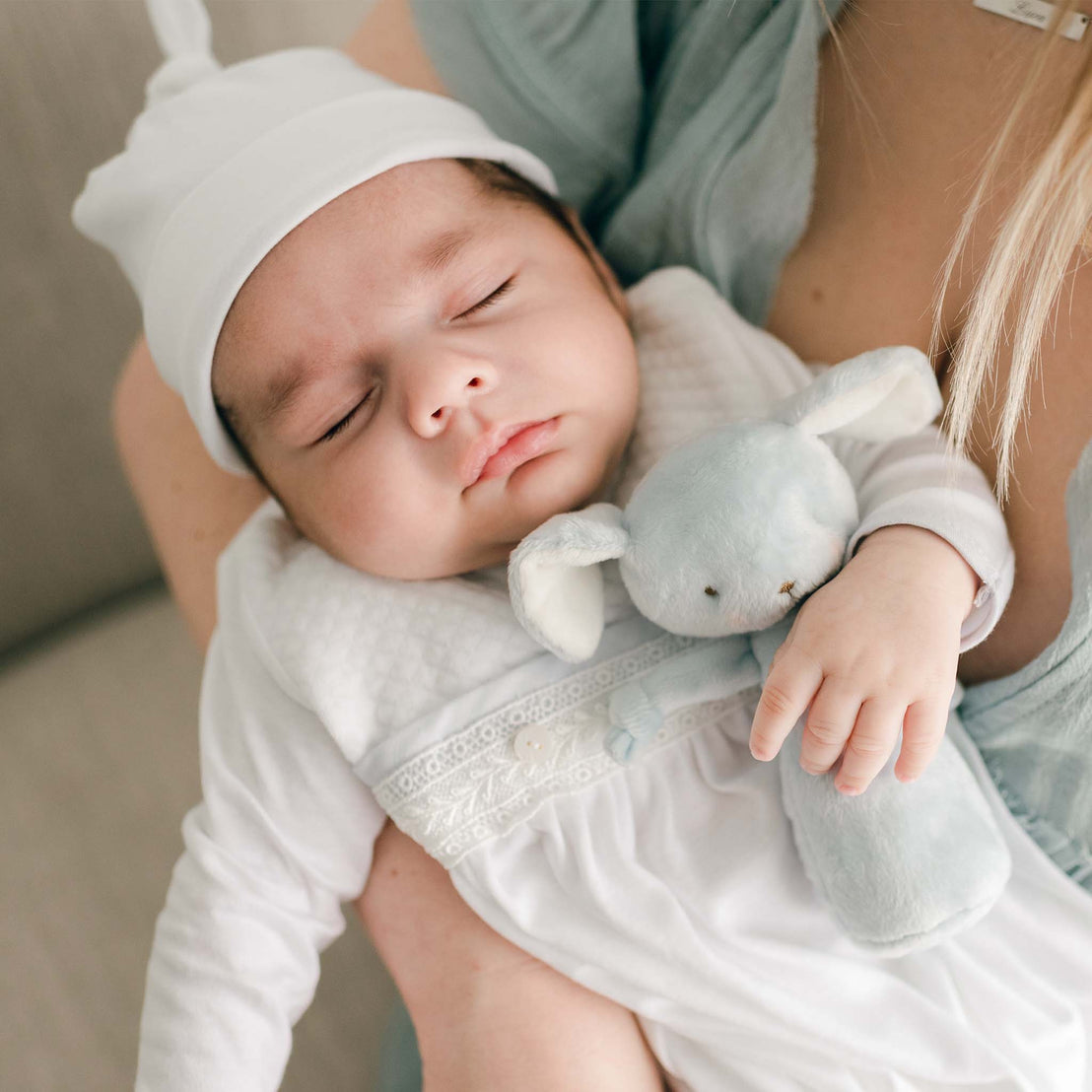 A close-up of a sleeping baby holding a soft blue plush bunny while wearing the Harrison Newborn Gown, with lace details and a textured finish.