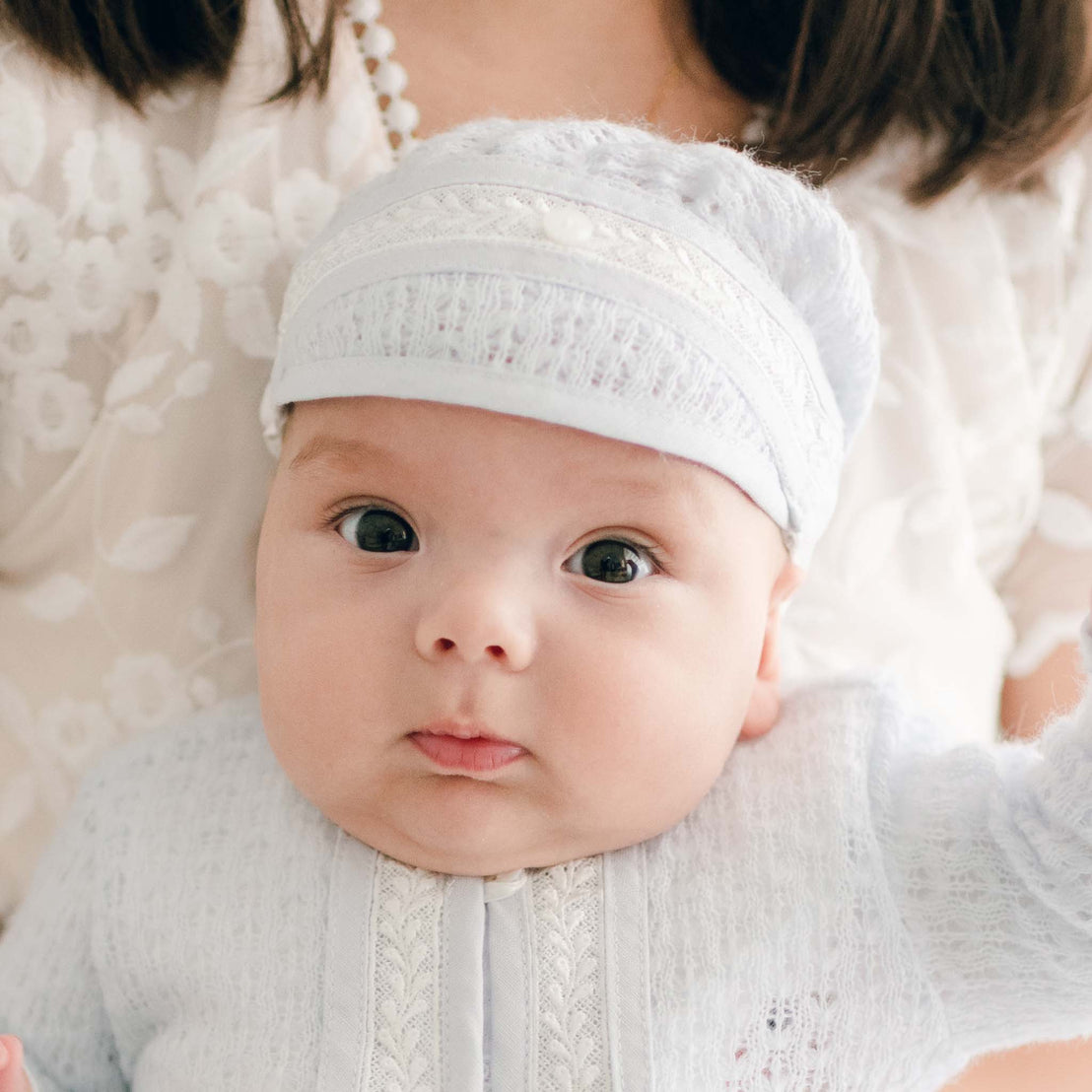 Close-up of a baby wearing the Harrison Newborn Knit Hat and matching sweater, featuring intricate lace knitting and delicate ivory lace trim with a pearl button on the front.