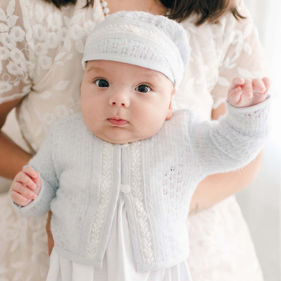 A baby wearing the Harrison Newborn Sweater, a soft light blue knit sweater with lace trim and button closures, over a white outfit.