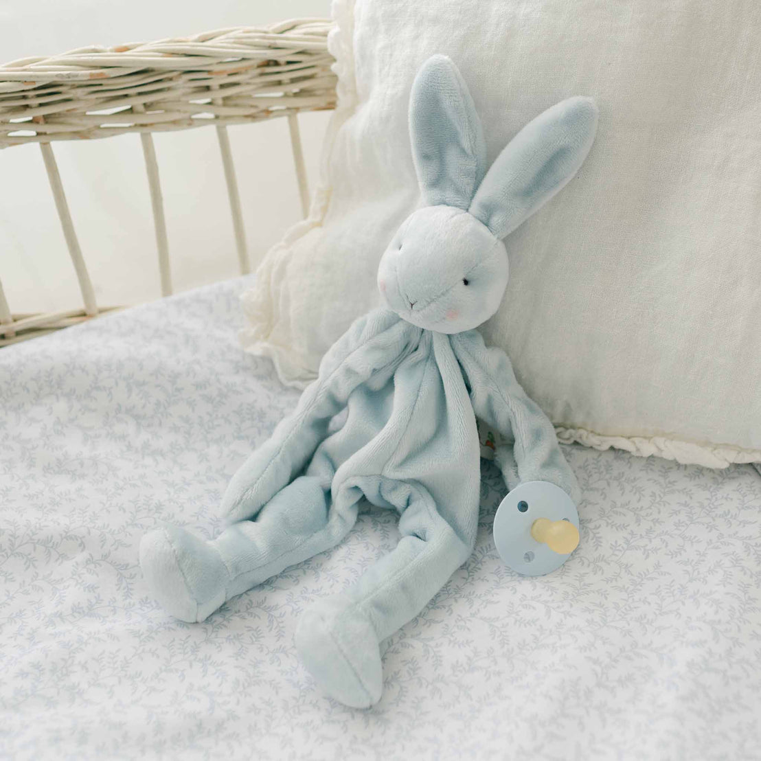 A close-up view of the Harrison Silly Bunny Buddy, a light blue plush bunny pacifier holder, resting on a soft floral-patterned surface with a neutral pillow in the background.