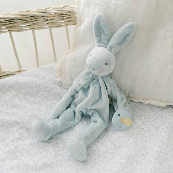 A close-up view of the Harrison Silly Bunny Buddy, a light blue plush bunny pacifier holder, resting on a soft floral-patterned surface with a neutral pillow in the background.