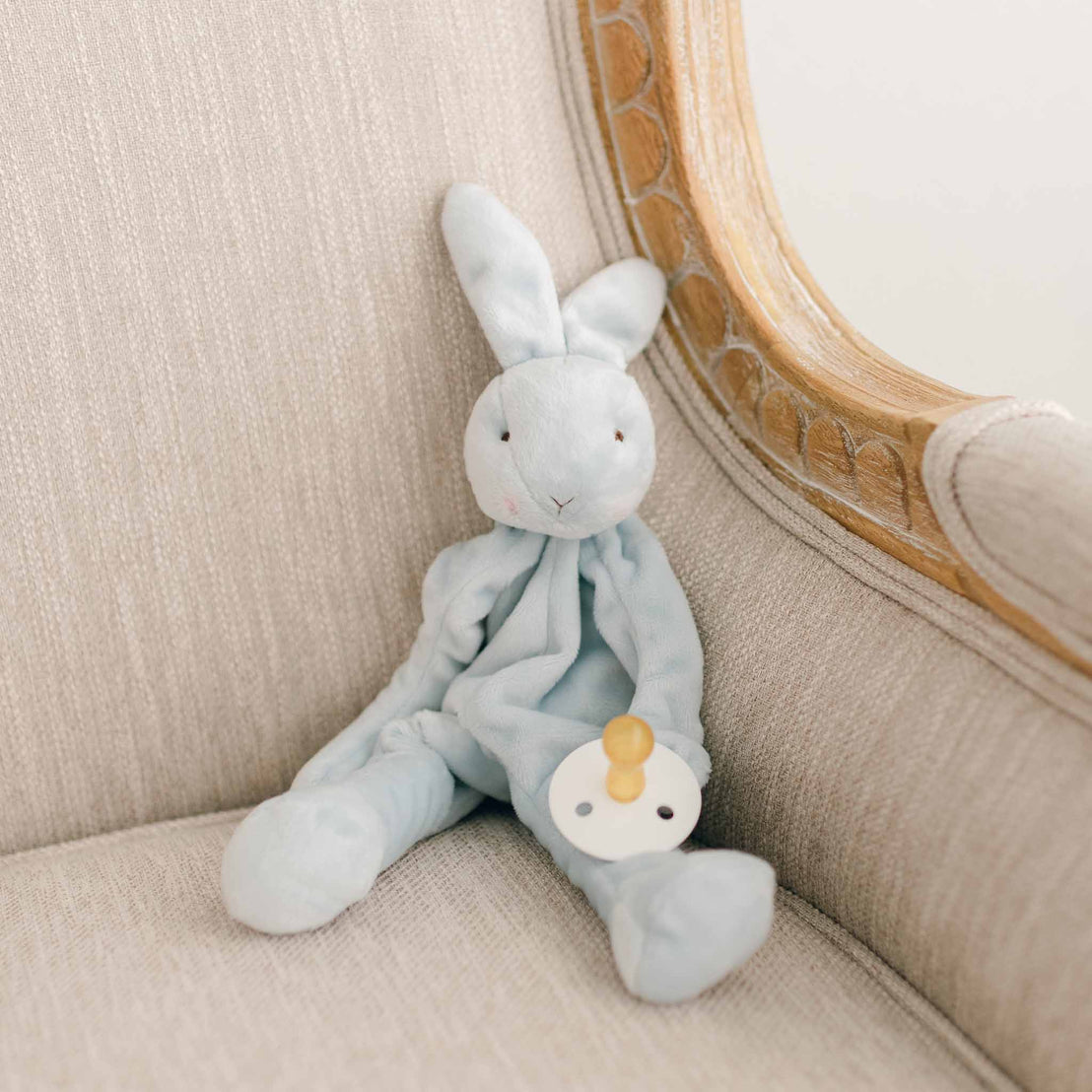 The Harrison Silly Bunny Buddy, a soft light blue plush pacifier holder, placed on a beige upholstered chair with its pacifier visible.