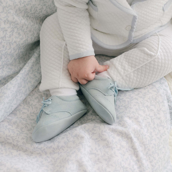 Close-up of a baby wearing the Harrison Suede Shoes with a soft blue suede exterior, delicate laces, and subtle perforated details, paired with a white quilted outfit.