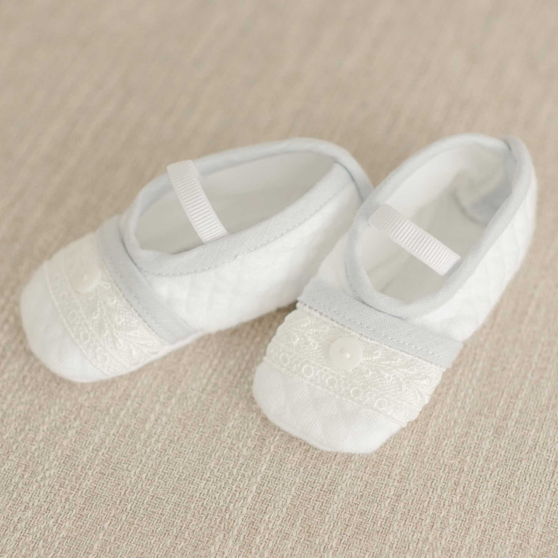 Harrison Booties displayed on a beige fabric background, showcasing lace accents and a button embellishment for an elegant baby look.