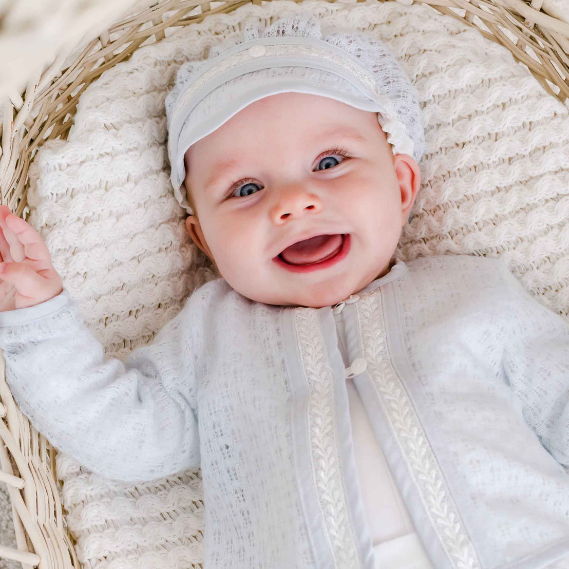 A happy baby lying in a woven basket, dressed in a delicate blue knit sweater with a lace-trimmed front, complemented by a matching outfit.