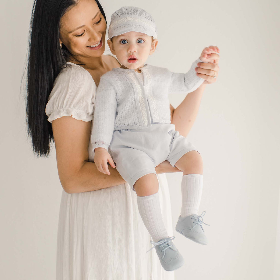 A baby wearing a soft blue knit sweater with intricate lace trim and matching shorts, held by a smiling woman in a light dress.
