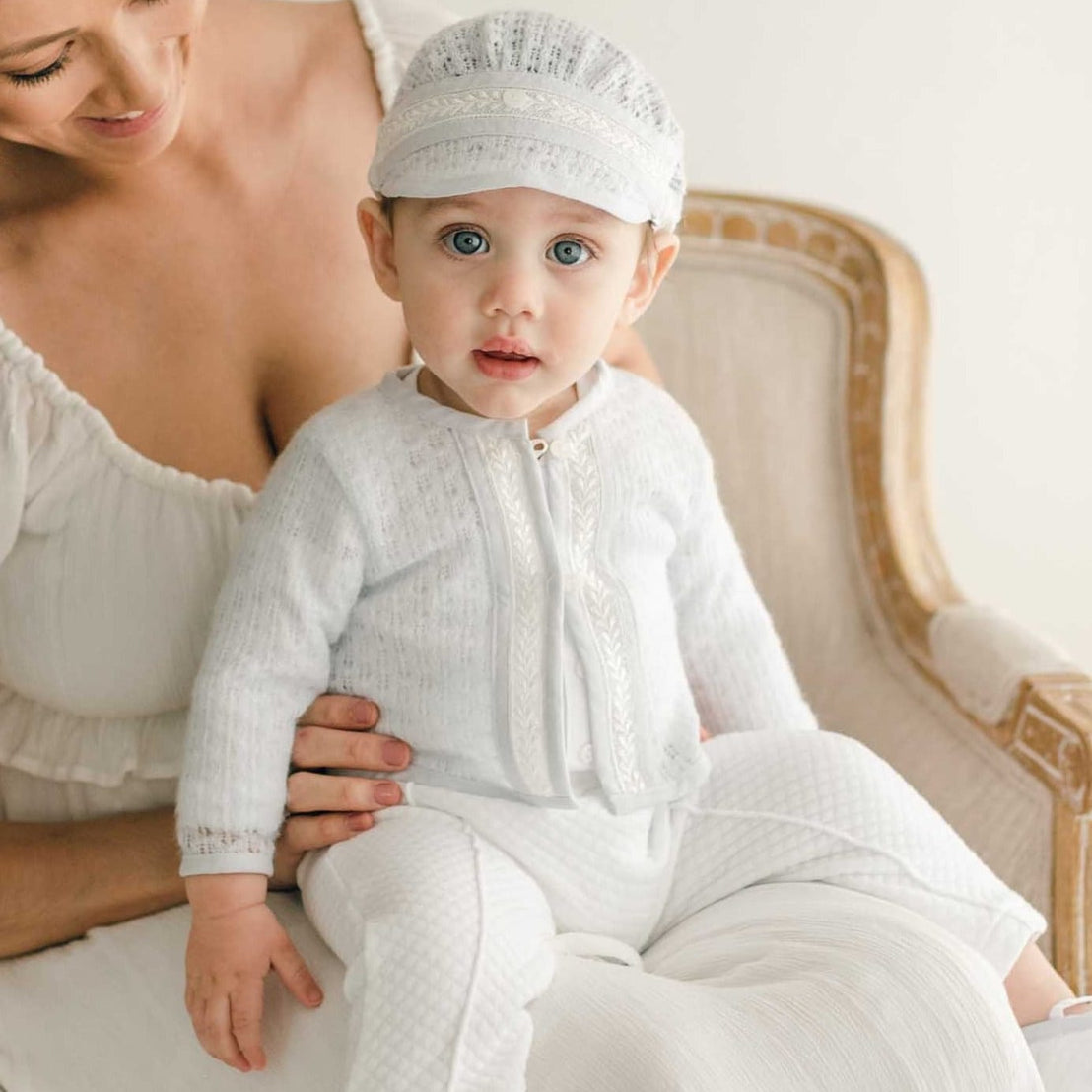 A baby sitting on his mother's lap wearing the Harrison Sweater 3-Piece Pant Suit, featuring a blue knit sweater with lace detailing, quilted white pants, and a matching knit cap.