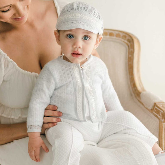 A baby sitting on his mother's lap wearing the Harrison Sweater 3-Piece Pant Suit, featuring a blue knit sweater with lace detailing, quilted white pants, and a matching knit cap.