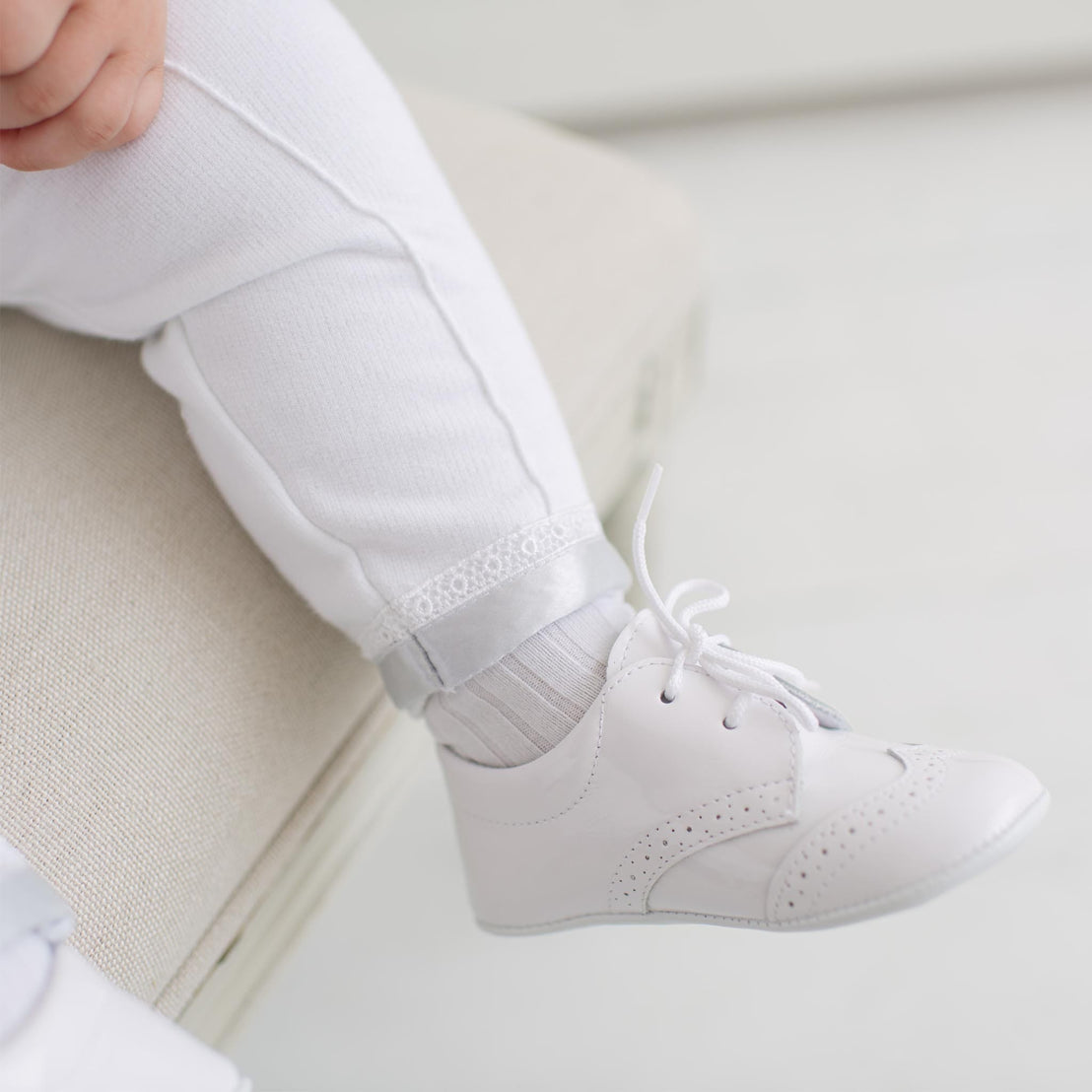 White shoes with laces worn by a baby sitting on a light-colored couch.
