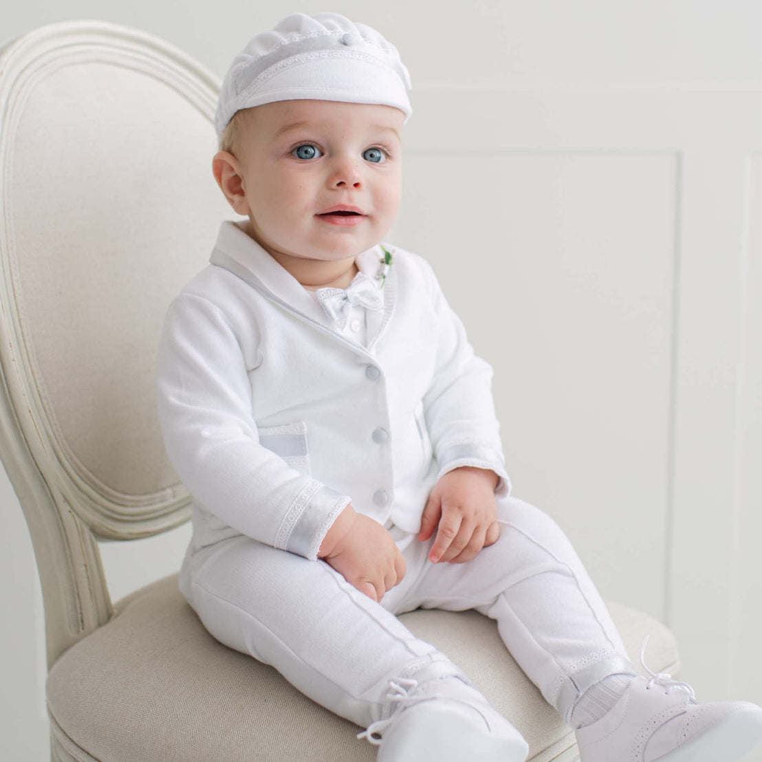 Baby in all white outfit sitting on a chair with a plain background.