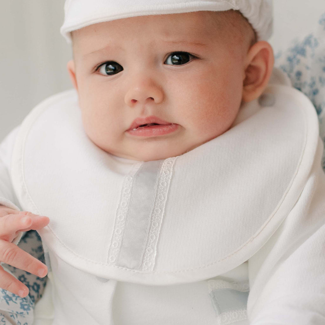 Baby in the Henry Brim Cap and matching 3-Piece Suit and Bib sitting on a woman with blue patterned dress. Part of Baby Beau & Belle's high end baby clothes.